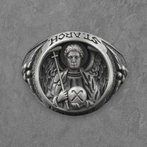 Puede incluir: Anillo plateado con un relieve detallado de un &aacute;ngel sosteniendo una cruz y un orbe. La banda del anillo est&aacute; inscrita con "ST. ARCH" en un patr&oacute;n circular. El dise&ntilde;o incluye detalles florales en los lados, creando un accesorio cl&aacute;sico de tem&aacute;tica religiosa.