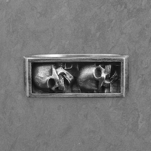 May include: A rectangular, silver-toned metal object featuring a detailed relief of two skulls. The skulls are positioned side-by-side within a rectangular frame, with visible teeth and eye sockets. The background is a solid, gray surface.