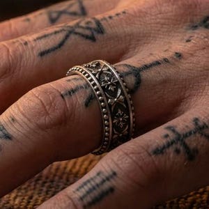May include: A silver ring featuring a detailed band with a repeating cross pattern and beaded edges. The ring is worn on a finger adorned with multiple black ink tattoos, showcasing a textured metallic finish and intricate craftsmanship.
