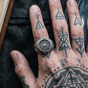 May include: A silver ring featuring a circular sunburst design on the face, worn on a finger. The ring has a textured, oxidized finish with intricate detailing around the central sunburst motif, creating a bold and distinct appearance.