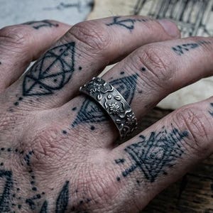 May include: A silver-toned metal ring featuring a detailed, embossed floral and leaf pattern across its entire outer surface. The band has a textured, oxidized finish that highlights the intricate botanical engravings throughout the metal.