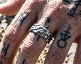 May include: A silver ring with a textured, uneven surface resembling rough stone or craggy rock. The band has a dark, oxidized finish that highlights the deep crevices and raised ridges across its entire exterior circumference.