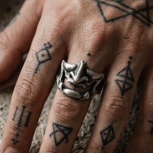 May include: A silver-toned ring featuring a sculpted mask design with prominent cheekbones and a defined nose bridge. The ring has a metallic finish and is worn on the middle finger, showcasing a detailed, stylized facial expression.