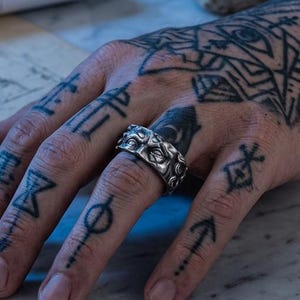 May include: A silver band ring featuring a repeating embossed pattern of stylized eyes. The ring has a dark, oxidized finish that highlights the intricate details of the eye motifs encircling the entire outer surface of the metal band.