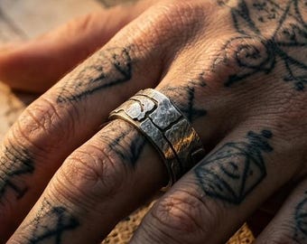 May include: A wide silver band ring featuring a textured, block-like surface pattern. The metal has a matte, weathered finish with irregular indentations that create a segmented appearance around the entire circumference of the ring.
