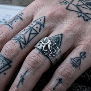 May include: A silver ring featuring a detailed octopus design with multiple tentacles wrapping around the band. The ring has a weathered, oxidized finish that highlights the intricate texture of the octopus, creating a distinct contrast against the metal surface.
