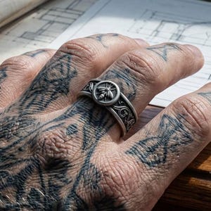 May include: A silver-toned metal ring featuring a circular centerpiece with a raised bee emblem, surrounded by an intricate engraved scrollwork pattern along the band. The ring is worn on a finger, showcasing the detailed metalwork and finish.
