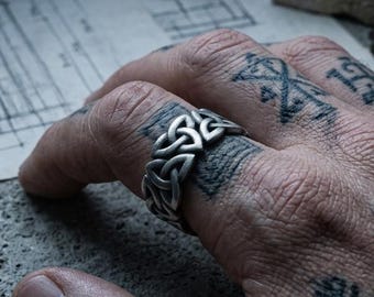 May include: A silver-toned metal ring featuring a repeating triquetra knot pattern. The band has a textured, oxidized finish that highlights the intricate, interlocking triangular shapes of the Celtic-inspired design throughout the entire circumference.
