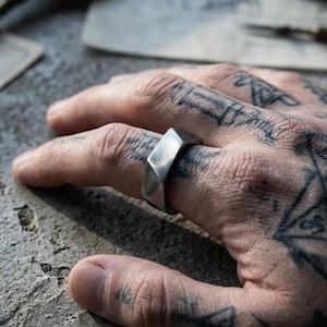 Könnte beinhalten: Ein silberfarbener Ring mit einer facettierten, geometrischen Oberflächenstruktur. Das Band weist mehrere flache, abgewinkelte Ebenen auf, die ein scharfes, mehrseitiges Erscheinungsbild erzeugen. Das Metall hat ein gebürstetes, mattes Finish.