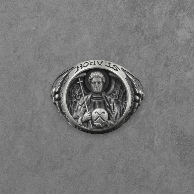 Puede incluir: Anillo plateado con un relieve detallado de un &aacute;ngel sosteniendo una cruz y un orbe. La banda del anillo est&aacute; inscrita con "ST. ARCH" en un patr&oacute;n circular. El dise&ntilde;o incluye detalles florales en los lados, creando un accesorio cl&aacute;sico de tem&aacute;tica religiosa.