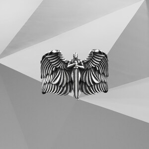 May include: A silver ring depicting an angel with spread wings and a sword. The ring has detailed feather textures and a central figure holding the sword. The background is a geometric pattern in shades of grey and white.