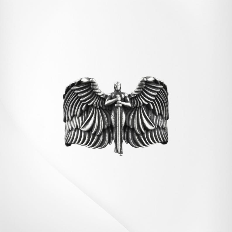 May include: A silver ring depicting an angel with spread wings and a sword. The ring has detailed feather textures and a central figure holding the sword. The ring is set against a white background.