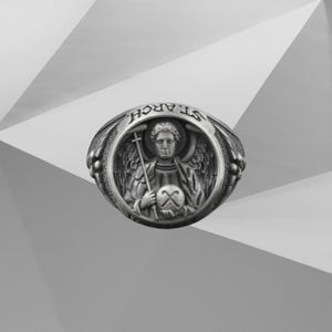 Puede incluir: Anillo plateado con la inscripci&oacute;n "ST. ARCH." grabada en la parte superior. El anillo presenta un relieve detallado de un &aacute;ngel sosteniendo una cruz y un globo. El &aacute;ngel tiene alas y est&aacute; rodeado por un marco circular.