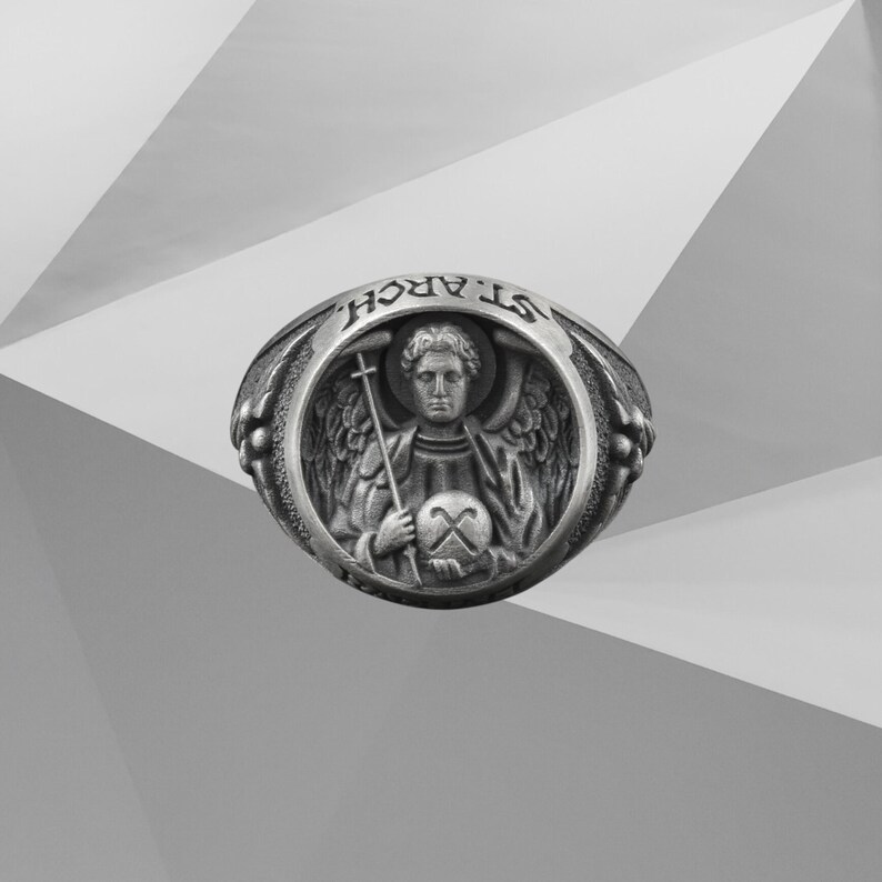 Puede incluir: Anillo plateado con la inscripci&oacute;n "ST. ARCH." grabada en la parte superior. El anillo presenta un relieve detallado de un &aacute;ngel sosteniendo una cruz y un globo. El &aacute;ngel tiene alas y est&aacute; rodeado por un marco circular.