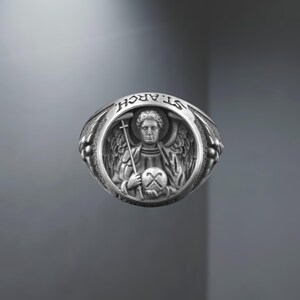 Puede incluir: Un anillo de plata con una detallada representaci&oacute;n de un &aacute;ngel sosteniendo una cruz y un orbe. El anillo est&aacute; inscrito con el texto "ST. ARCH." en la parte superior. El anillo tiene un estilo vintage detallado.