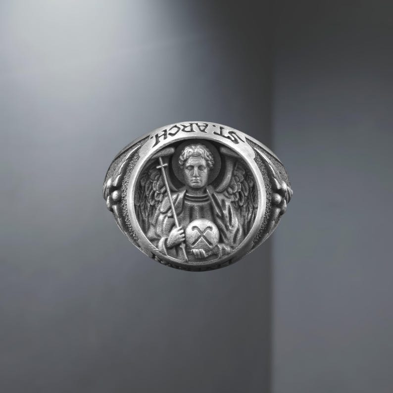 Puede incluir: Un anillo de plata con una detallada representaci&oacute;n de un &aacute;ngel sosteniendo una cruz y un orbe. El anillo est&aacute; inscrito con el texto "ST. ARCH." en la parte superior. El anillo tiene un estilo vintage detallado.