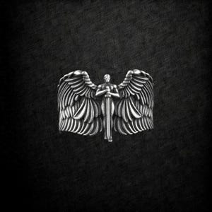 May include: A silver ring depicting an angel with wings and a sword. The ring's design shows the angel holding the sword, with detailed feathering on the wings. The background is dark, highlighting the ring's intricate details.