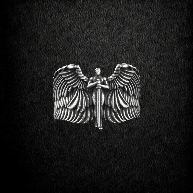 May include: A silver ring depicting an angel with wings and a sword. The ring's design shows the angel holding the sword, with detailed feathering on the wings. The background is dark, highlighting the ring's intricate details.