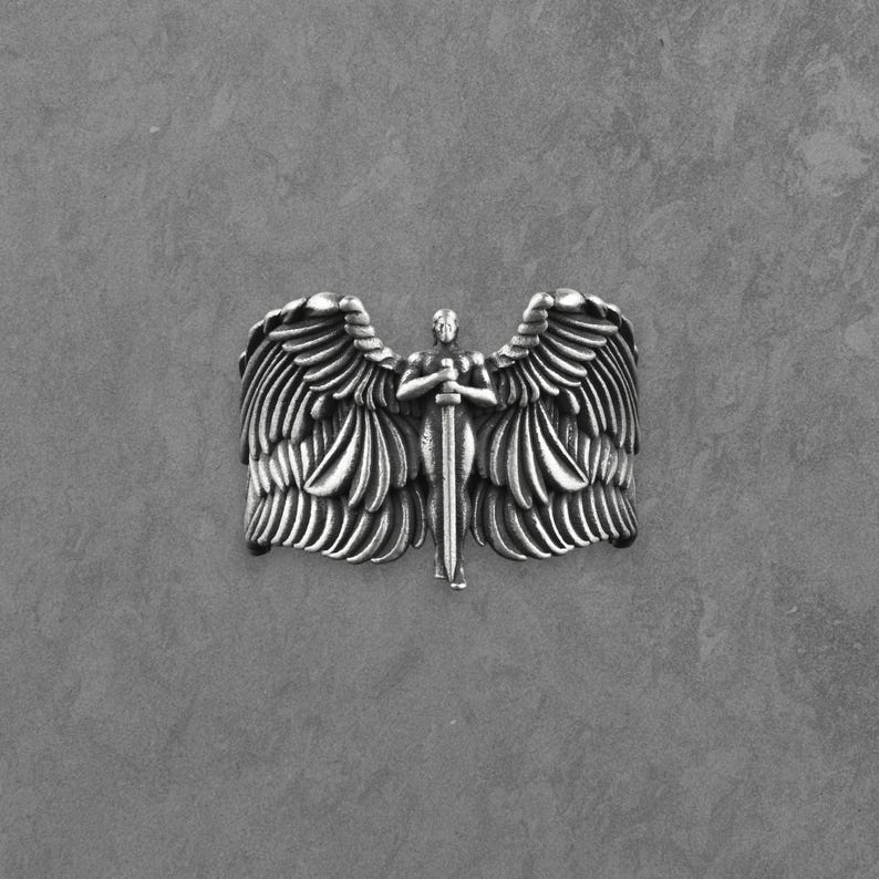 May include: A silver ring depicting an angel with large, detailed wings and a sword. The angel is the central focus, with intricate feather detailing. The ring has a textured, metallic appearance.