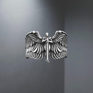May include: A silver ring depicting an angel with wings and a sword. The ring has detailed feather and wing designs, and the angel figure is centered. The ring is a statement piece, suitable for everyday wear.