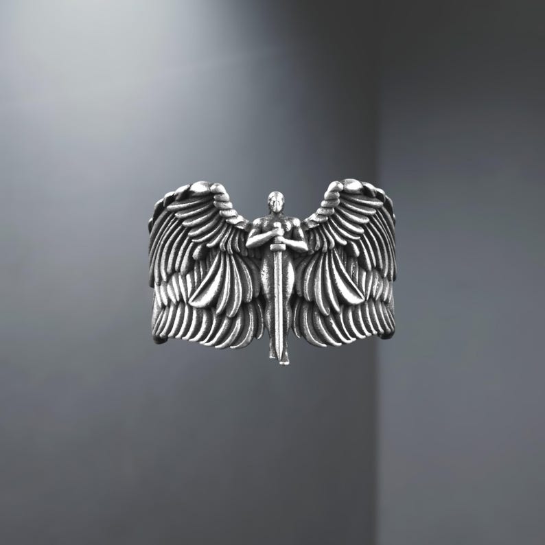 May include: A silver ring depicting an angel with wings and a sword. The ring has detailed feather and wing designs, and the angel figure is centered. The ring is a statement piece, suitable for everyday wear.