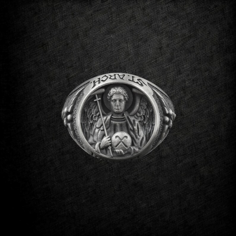 Puede incluir: Anillo de plata con un relieve detallado de un &aacute;ngel sosteniendo una cruz y un globo. El anillo est&aacute; inscrito con las palabras "ST. ARCH." alrededor del &aacute;ngel. El anillo tiene un dise&ntilde;o detallado y texturizado sobre un fondo oscuro.
