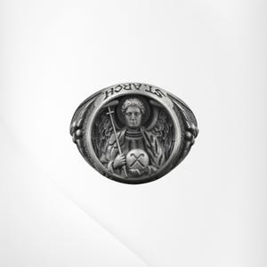 Puede incluir: Anillo plateado con una detallada representaci&oacute;n de un &aacute;ngel sosteniendo una cruz y un globo. El anillo est&aacute; grabado con las palabras "ST. ARCH." alrededor de la imagen central. Dise&ntilde;o detallado y texturizado.