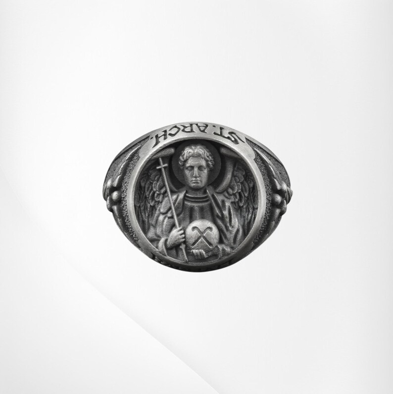 Puede incluir: Anillo plateado con una detallada representaci&oacute;n de un &aacute;ngel sosteniendo una cruz y un globo. El anillo est&aacute; grabado con las palabras "ST. ARCH." alrededor de la imagen central. Dise&ntilde;o detallado y texturizado.