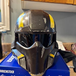 Op de afbeelding: Een verweerde, zwart-gele helm met een vizier. De helm heeft een versleten uiterlijk met zichtbare slijtage. Het vizier is donker en de helm ligt op een blauwe doos.