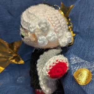 May include: Crocheted doll with a black and white outfit, red accents, and a gold crown keychain. The doll has a light pink face and white hair. The keychain is attached to the top of the doll's head. The doll is handmade.