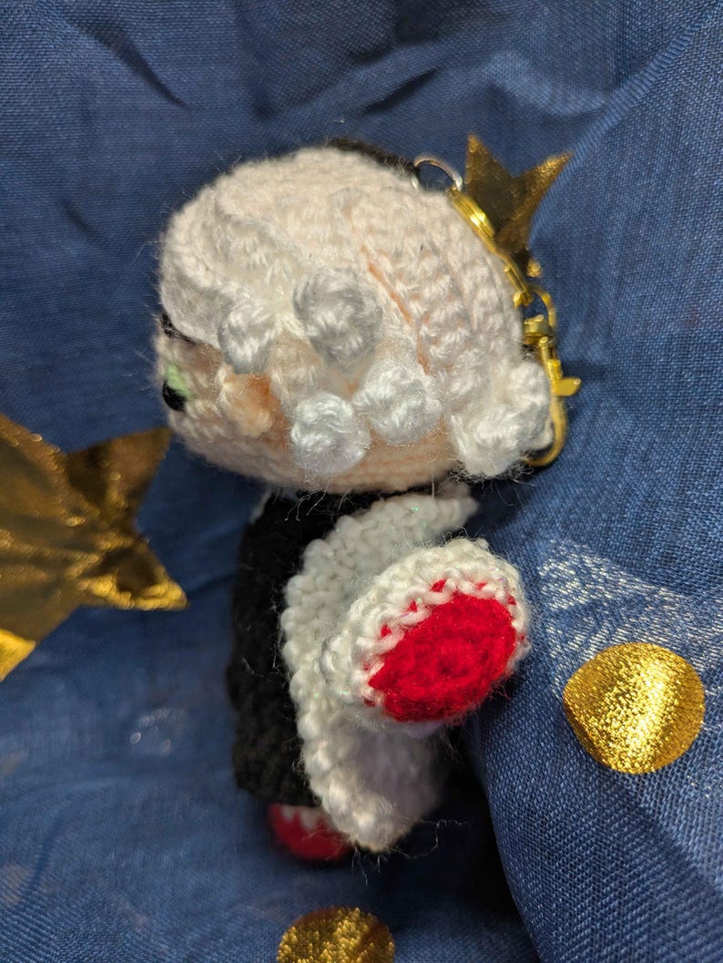 May include: Crocheted doll with a black and white outfit, red accents, and a gold crown keychain. The doll has a light pink face and white hair. The keychain is attached to the top of the doll's head. The doll is handmade.