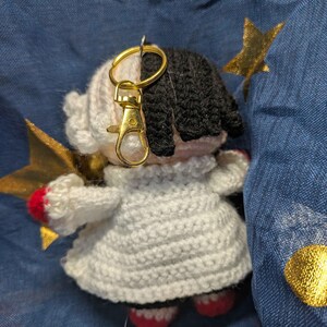 May include: A handmade crochet doll keychain with a gold-colored clasp. The doll has black and white hair, a white dress, and red accents on the feet and hands. A gold star is attached to the doll.