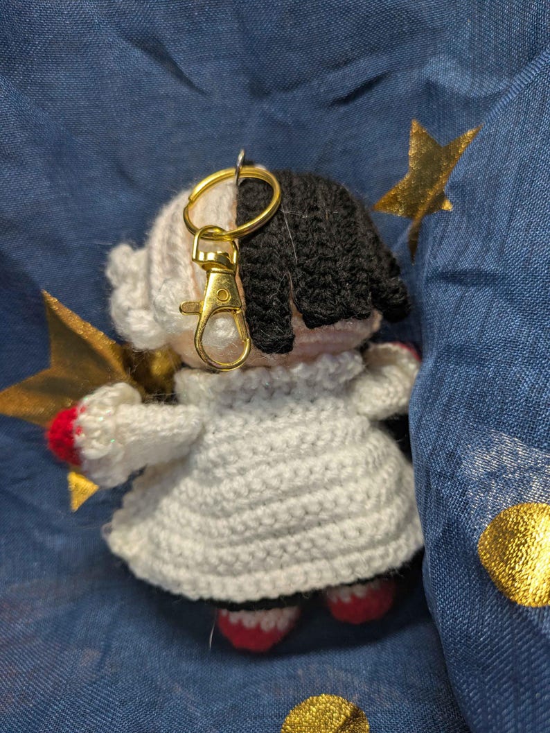 May include: A handmade crochet doll keychain with a gold-colored clasp. The doll has black and white hair, a white dress, and red accents on the feet and hands. A gold star is attached to the doll.