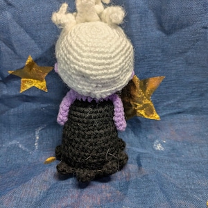 May include: A handmade crochet doll featuring a white head with curly hair, a purple collar and arms, and a black dress. The doll is set against a blue background with gold star and circle accents. The doll is a handmade craft.