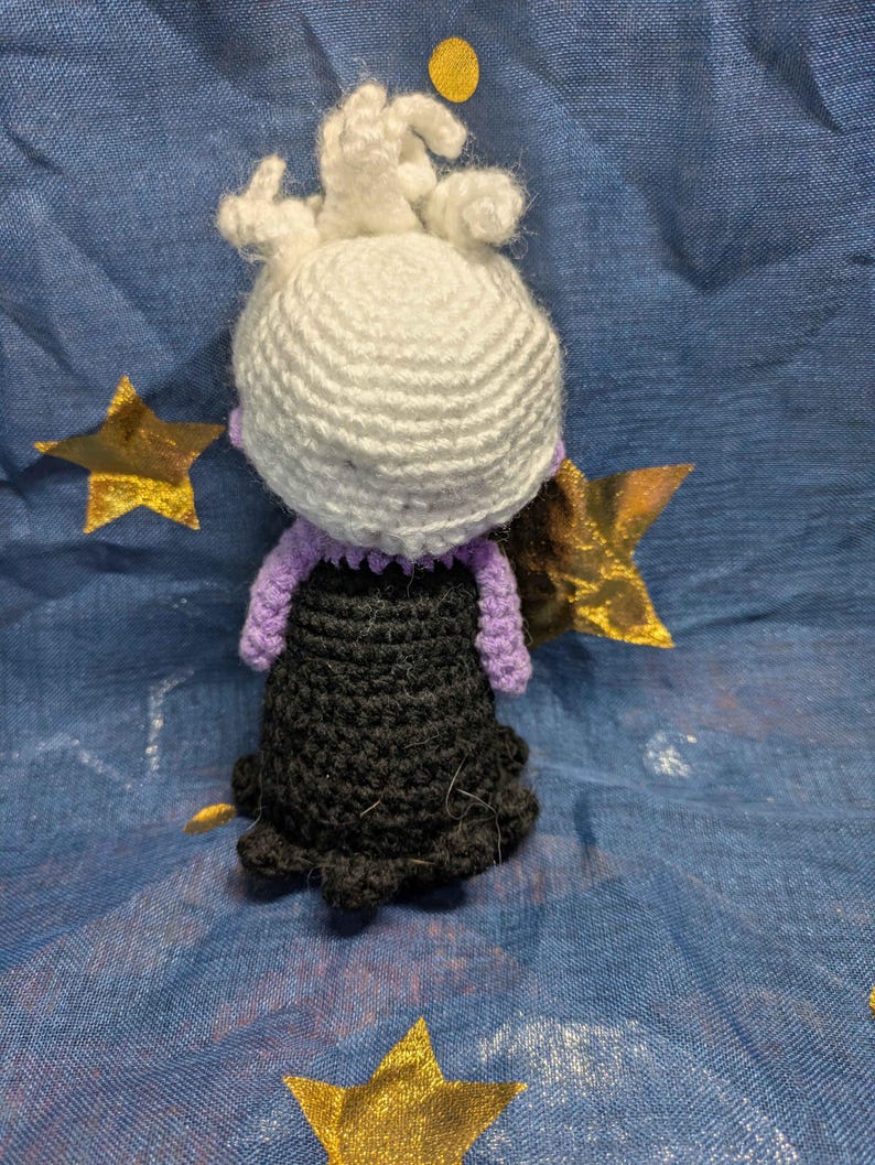 May include: A handmade crochet doll featuring a white head with curly hair, a purple collar and arms, and a black dress. The doll is set against a blue background with gold star and circle accents. The doll is a handmade craft.