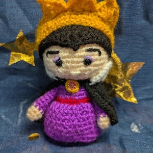 May include: A handmade crochet doll of a character wearing a purple dress, black cape, and a yellow crown. The doll has a white face with black hair and purple eye shadow. A red belt and gold embellishments complete the look. The doll is approximately 4 inches tall.