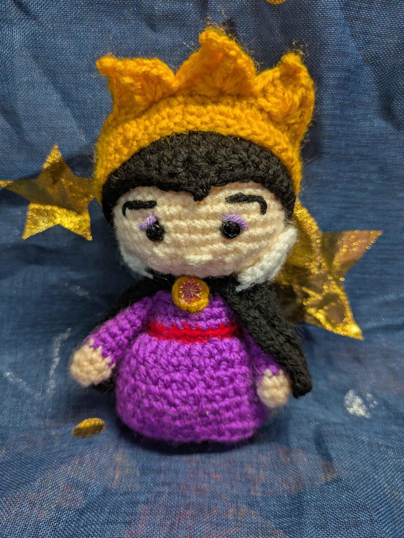 May include: A handmade crochet doll of a character wearing a purple dress, black cape, and a yellow crown. The doll has a white face with black hair and purple eye shadow. A red belt and gold embellishments complete the look. The doll is approximately 4 inches tall.