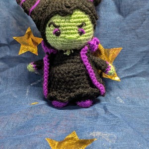 May include: A handmade crochet doll of a character with a green face, black hair, and a black dress with purple accents. The doll has horns and is posed on a blue fabric with gold star accents. The doll is approximately 5 inches tall.