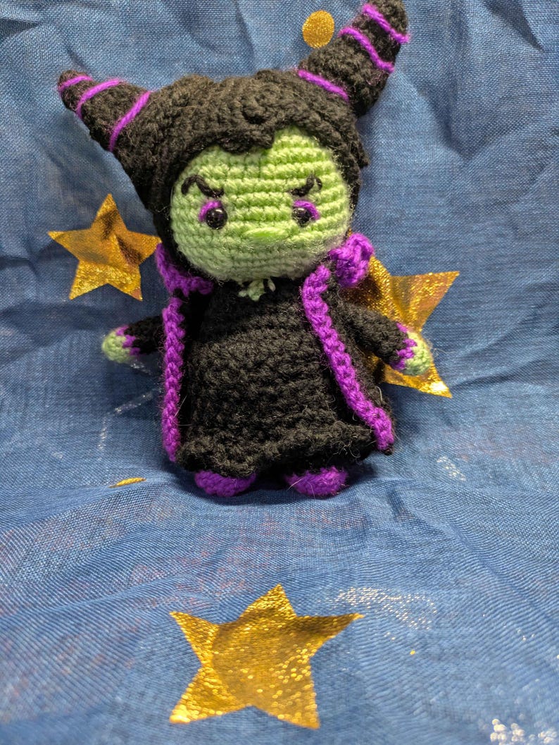 May include: A handmade crochet doll of a character with a green face, black hair, and a black dress with purple accents. The doll has horns and is posed on a blue fabric with gold star accents. The doll is approximately 5 inches tall.