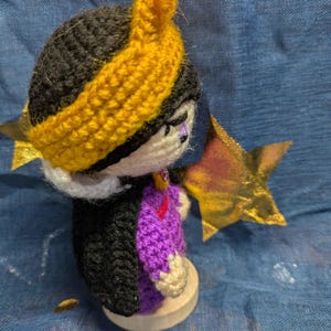 May include: A handmade crochet doll with a black head and a yellow headband. The doll wears a purple and black outfit and has a gold star accessory. The doll is on a wooden base. The doll is a handmade craft.