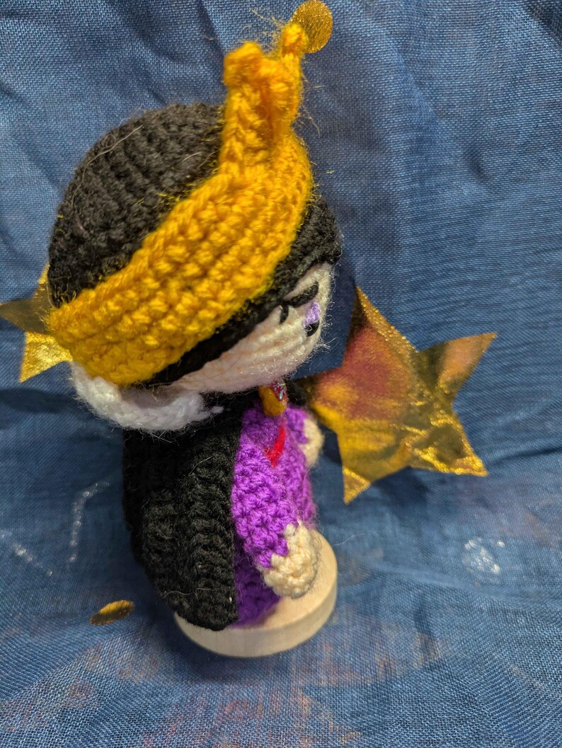 May include: A handmade crochet doll with a black head and a yellow headband. The doll wears a purple and black outfit and has a gold star accessory. The doll is on a wooden base. The doll is a handmade craft.