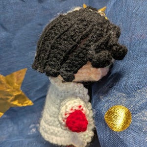 May include: Crocheted doll with black hair, a white cape with red accents, and a small gold crown. The doll is handmade and appears to be a character from a story or game. The doll is set against a blue background with gold star and circle decorations.