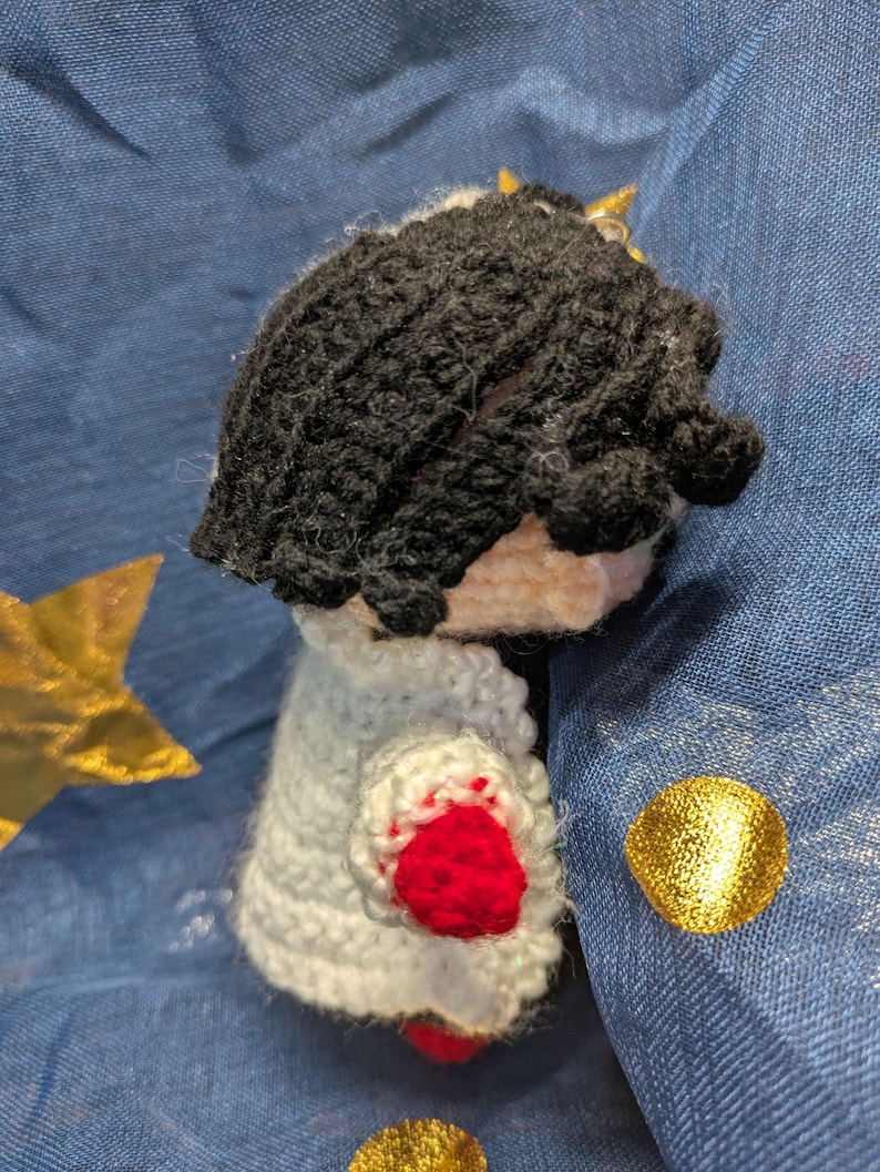 May include: Crocheted doll with black hair, a white cape with red accents, and a small gold crown. The doll is handmade and appears to be a character from a story or game. The doll is set against a blue background with gold star and circle decorations.