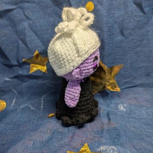 May include: A handmade crochet doll with a white bonnet, purple face, and black dress. The doll is set against a blue background with gold star and circle accents. The doll's features are detailed with black thread.