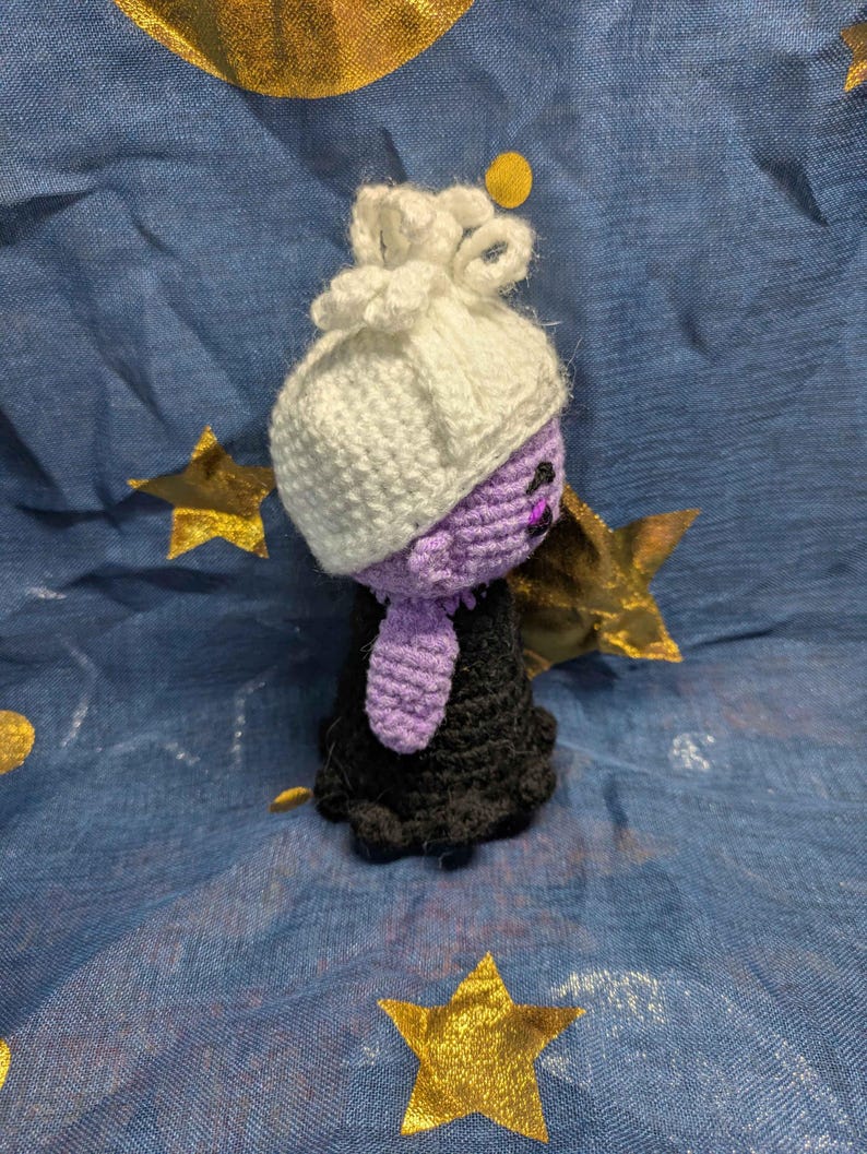 May include: A handmade crochet doll with a white bonnet, purple face, and black dress. The doll is set against a blue background with gold star and circle accents. The doll's features are detailed with black thread.