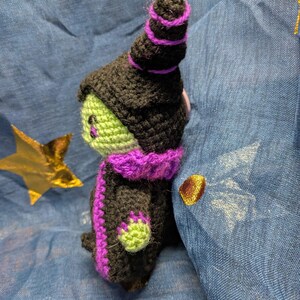 May include: A handmade crochet witch doll with a black hat and robe, accented with purple trim. The witch has a green face and hands. The doll is set against a blue background with gold star accents.