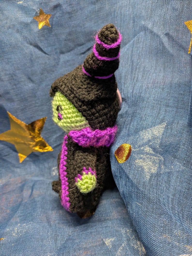 May include: A handmade crochet witch doll with a black hat and robe, accented with purple trim. The witch has a green face and hands. The doll is set against a blue background with gold star accents.