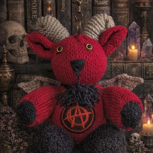 Pode incluir: Um brinquedo de pelúcia vermelho tricotado com chifres, asas e uma gola preta. O brinquedo tem um símbolo bordado em preto e vermelho no peito. O fundo inclui livros antigos, velas e cristais, criando um tema gótico.