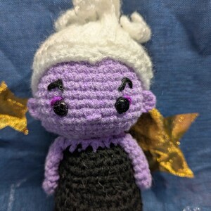 May include: A handmade crochet doll of a character with purple skin, black eyes, and a white headpiece. The doll is wearing a black dress. Gold star-shaped decorations are on either side of the doll.