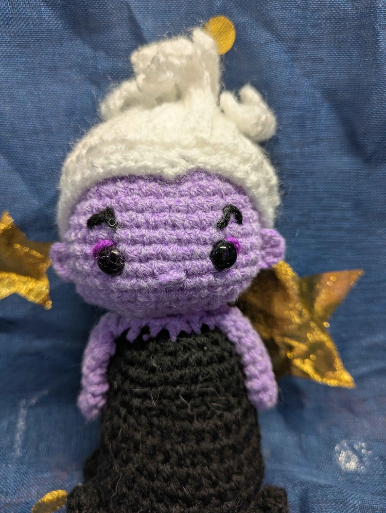 May include: A handmade crochet doll of a character with purple skin, black eyes, and a white headpiece. The doll is wearing a black dress. Gold star-shaped decorations are on either side of the doll.